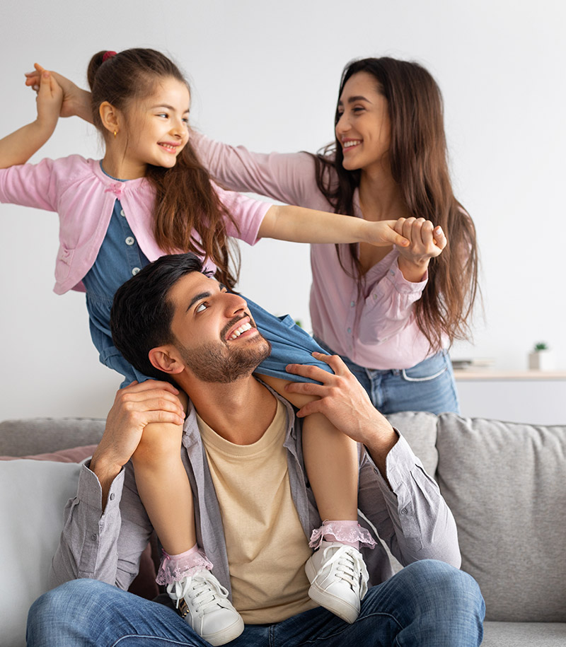 Smiling family at home, with a father sitting on a couch while a young daughter sits on his shoulders and a mother holds the child’s hand.
