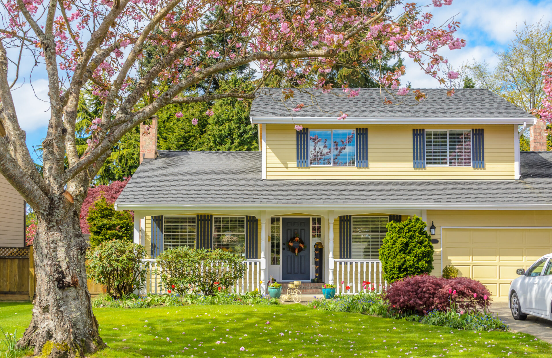 Yellow two-story suburban house with a gray roof, front porch, and manicured lawn, framed by a blooming pink cherry blossom tree in spring.